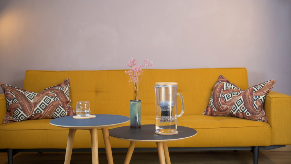 The Lotus Vita ENYA water filter pitcher on a coffee table in a modern living room, with a glass of filtered water nearby.