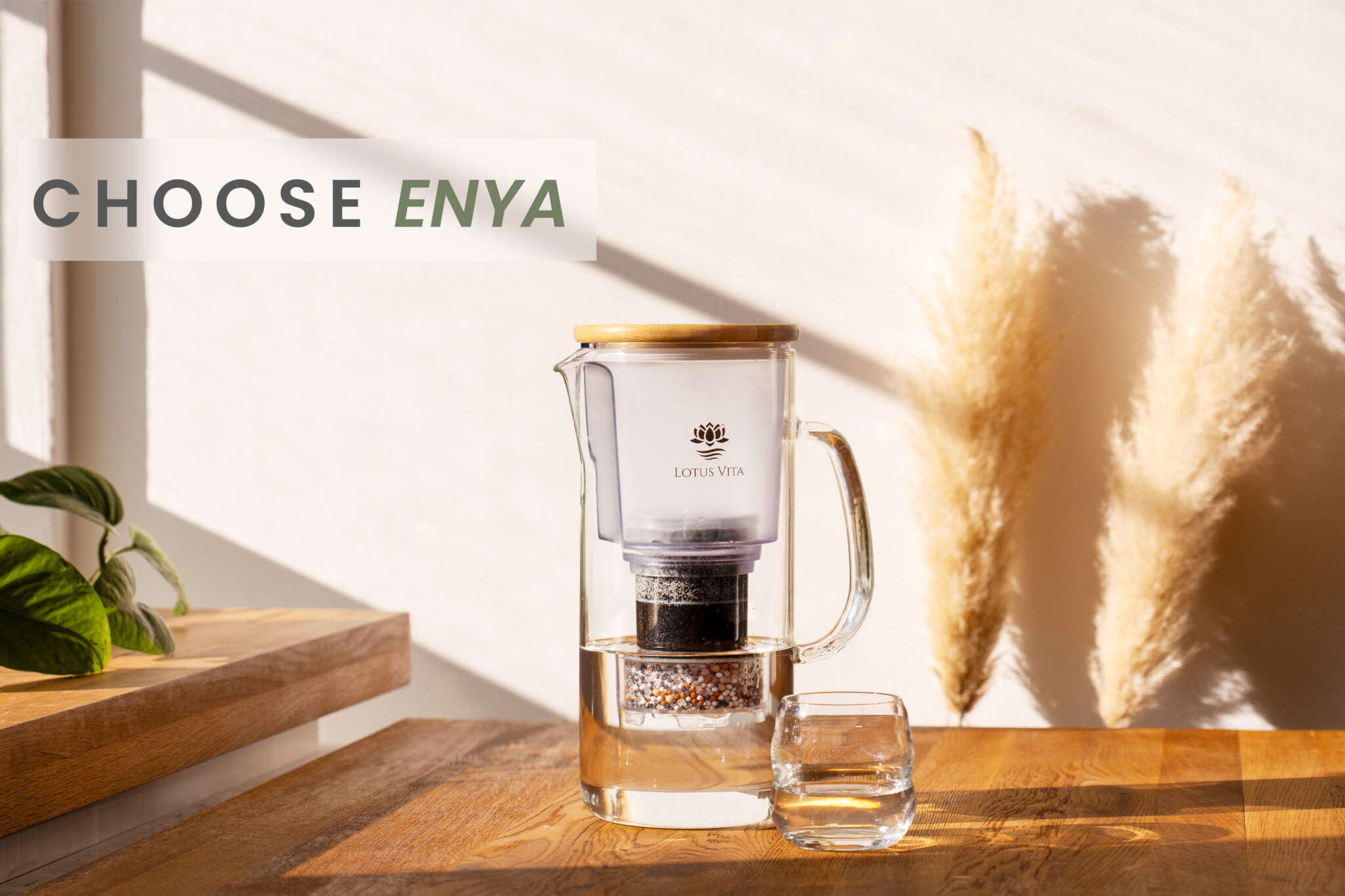 The Lotus Vita ENYA glass water filter pitcher with a bamboo lid, sitting on a wooden table in the sunlight next to a filled glass of water.