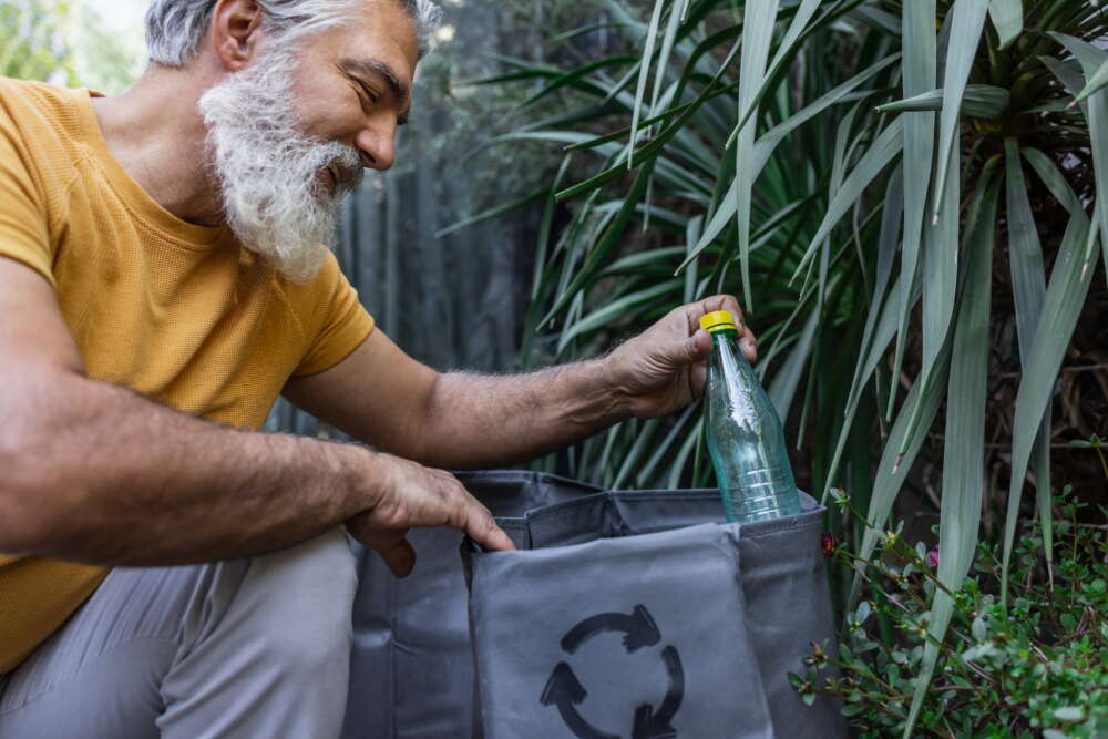 A smiling man with a white beard places a plastic bottle into a recycling bag, embodying a commitment to environmental sustainability.
